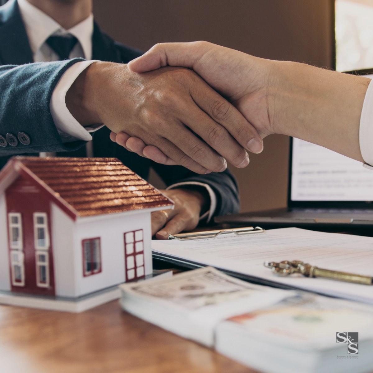 A handshake across a desk over a signed property contract, with house keys and a model house beside it.